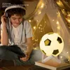A young boy wearing headphones sits on the floor in a tent decorated with fairy lights, looking at his phone. A soccer ball lamp is on a stand next to him. Produktbild-Vorschau