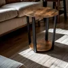 A round side table with a wood-look top and black metal legs is placed next to a sofa on a rug. Sunlight streams in, creating shadows. Podgląd zdjęcia produktu
