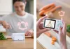 A split image showing a baby monitor in use. The left side shows a woman preparing food in a kitchen with the baby monitor on the counter. The right side shows hands holding the baby monitor displaying a baby. Both sides have a bell icon overlay. Náhľad obrázku produktu