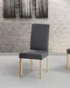 A single gray upholstered dining chair with wooden legs is shown in a room with a brick wall and artwork. Náhled obrázku produktu