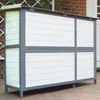 A wooden animal hutch with white and gray panels, standing on legs on a brick patio. Produktbild-Vorschau