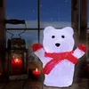 A festive polar bear decoration sits on a wooden surface in front of a window with a snowy scene outside. Two red candles are lit, one in a lantern and one beside it, creating a warm, cozy atmosphere. Anteprima dell’immagine prodotto