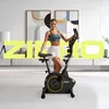 A woman stands next to a stationary bike in a modern living room. Produktbild-Vorschau