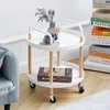 A white two-tier round rolling cart with wooden legs and handles, styled with a vase of flowers and a cup on the top shelf, and a book on the bottom shelf. The cart is placed on a wooden floor next to a gray armchair. A stack of books is visible in the bac Anteprima dell’immagine prodotto