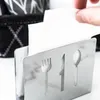 A hand is pulling a white napkin from a stainless steel napkin holder with cutouts of cutlery. Produktbild-Vorschau