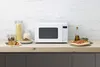 A white microwave oven is displayed on a kitchen counter, surrounded by food items like pasta, fried chicken, and pizza. Produktbild-Vorschau