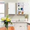 A white wall-mounted cabinet with an open door, displaying kitchen items, is situated above a kitchen counter with a sink and faucet. To the left, a closed white cabinet is mounted on the wall. In the foreground, a vase of yellow lilies sits on a wooden su Produktbild-Vorschau