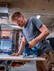 A man is using a jigsaw to cut a piece of wood in a workshop. Anteprima dell’immagine prodotto