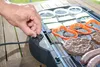 A hand is shown adjusting the temperature knob on an electric grill where burgers, sausages, and vegetables are cooking. Náhled obrázku produktu