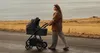 A woman pushing a stroller on a paved path near a body of water and dry grass. Produktbild-Vorschau