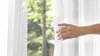 A hand is pulling back a sheer white curtain, revealing a blurred background of green trees and a building. Produktbild-Vorschau