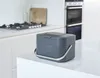 A dark gray compost bin with a silver handle sits on a white kitchen counter, with a stovetop and oven visible in the background. Produktbild-Vorschau