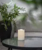 A white LED candle sits on a glass-topped table next to a potted plant. Produktbild-Vorschau
