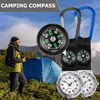 A person in outdoor gear stands in a grassy, mountainous area with a tent in the background. Several compass watches are displayed prominently in the foreground. Produktbild-Vorschau