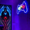 A neon gamepad sign is mounted on a brick wall in a room with purple lighting. A large digital artwork featuring a hooded figure is displayed on a screen to the left. Produktbild-Vorschau