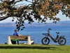 A person sits on a bench by a lake, listening to music, with an e-bike parked nearby. Produktbild-Vorschau