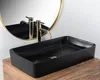 A black rectangular vessel sink with gold faucets is shown on a wooden vanity, with a gold-framed mirror in the background. Produktbild-Vorschau