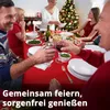 A family is celebrating Christmas around a table with a red tablecloth, food, and drinks. A Christmas tree is visible in the background. Produktbild-Vorschau