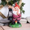 A garden gnome holding a lantern, standing on a grassy mound with a mushroom at its base, surrounded by rocks and wooden planks. Produktbild-Vorschau
