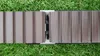 A close-up view of two composite deck boards joined by a silver mounting clip, resting on a green grassy surface. Produktbild-Vorschau