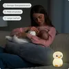 A young woman is breastfeeding a baby on a couch, with a small penguin-shaped night light on the couch next to them. Produktbild-Vorschau