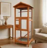 A wooden bird cage is shown in a living room setting, next to a sofa and a wooden table. Produktbild-Vorschau