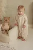 A baby is standing next to a child-sized armchair with a teddy bear sitting in it. Produktbild-Vorschau