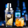 A cocktail shaker and a glass of cocktail are displayed on a dark wooden surface with a blurred background of bottles. Produktbild-Vorschau