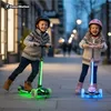 Two children riding illuminated scooters on a sidewalk at dusk. Anteprima dell’immagine prodotto