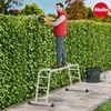 A man is trimming a hedge while standing on a multi-purpose ladder. A rake and a bag are nearby. Anteprima dell’immagine prodotto