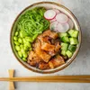 A close-up overhead view of a poke bowl with salmon, edamame, seaweed salad, cucumber, and radish slices. Chopsticks are visible in the foreground. Produktbild-Vorschau