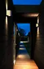 A walkway lined with dark wood walls and illuminated by wall-mounted lights, leading to an open outdoor space. Anteprima dell’immagine prodotto
