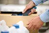 A person is using a utility knife to open a cardboard box containing plastic bottles with blue caps. Voir les images du produit