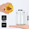 A hand holds a jar lid with a bee and flower design, while another hand holds a glass jar with dimensions indicated. Voir les images du produit