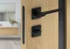 A close-up shot of a modern black door handle set on a light wood door, with a blurred background of a bathroom interior. Produktbild-Vorschau