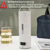 A white portable electric kettle is shown on a table next to a glass of liquid and a bag. Produktbild-Vorschau