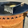 A close-up shot of a metal fireplace tool resting on a black grill surface, with a rusty metal fire pit in the foreground featuring flame cutouts. Podgląd zdjęcia produktu