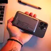 A hand holding a smartphone with a power bank attached, with a laptop and pen in the background. Produktbild-Vorschau