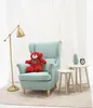 A teddy bear sits on a chair next to a lamp and side tables. Podgląd zdjęcia produktu