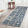A long runner rug with a tile pattern is laid out on a light wooden floor in a hallway. A white console table with books on it is to the left, and a wooden accent table is to the right. Produktbild-Vorschau