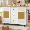 A white kitchen island with a wooden countertop is shown in a bright, airy room with large windows. The island has sliding doors with a rattan insert and drawers. A basket of books is on the floor in front of it. Produktbild-Vorschau