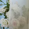 A close-up view of white roses behind a sheer, beige curtain with a floral pattern. Produktbild-Vorschau