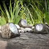 Three solar-powered rock lights are placed on a stone path next to tall green grass. Produktbild-Vorschau