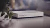 A close-up shot of a white smart home hub device placed on a table, with a plant in the foreground and a book in the background. Anteprima dell’immagine prodotto
