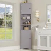 A tall, narrow gray corner cabinet with open shelves and two doors, placed in a bathroom setting next to a window. Náhled obrázku produktu