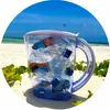 A clear pitcher filled with crumpled plastic bottles sits on a sandy beach with green foliage in the foreground and the ocean and sky in the background. Podgląd zdjęcia produktu