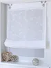 A white sheer Roman blind with embroidered floral pattern hangs in a window frame, with decorative heart-shaped objects on the windowsill below. Produktbild-Vorschau