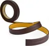 A roll of dark brown self-adhesive trim tape with a yellow adhesive backing is shown against a white background. The tape is partially unrolled, revealing its flexible nature and a subtle ridge detail. Náhľad obrázku produktu