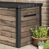 A wooden-look storage box with a black lid and frame, placed outdoors next to a brick wall, with potted plants in the foreground. Produktbild-Vorschau