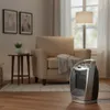 A ceramic heater is placed on a wooden floor in a living room, next to an armchair and a side table. Podgląd zdjęcia produktu